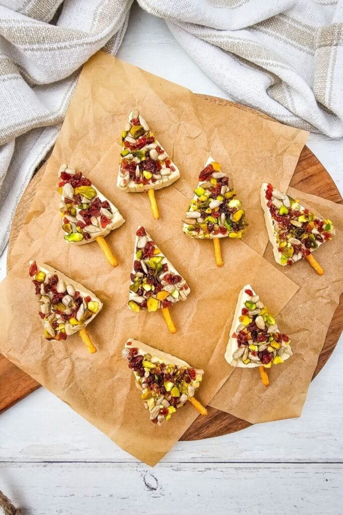 Christmas tree–shaped cream cheese slices with pretzel trunks, topped with chopped nuts and dried fruit, arranged on parchment over a wooden board.