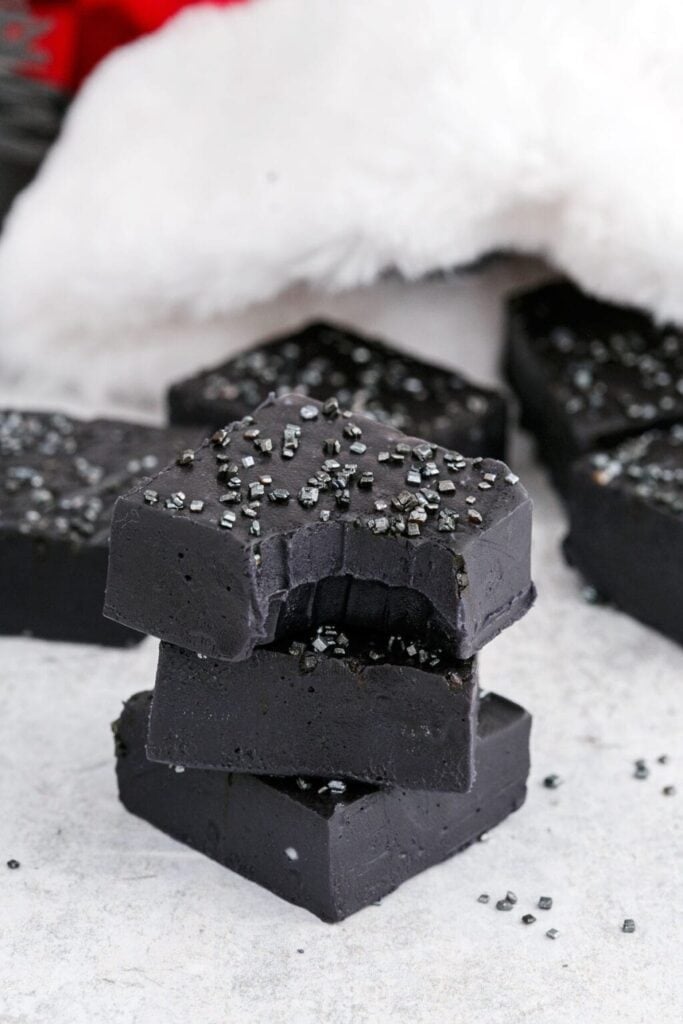 Black fudge squares topped with shiny sugar crystals, stacked on a light surface with soft white fabric in the background.