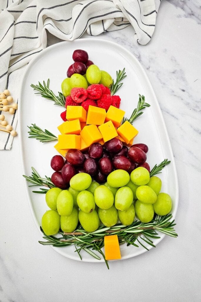 Christmas tree–shaped platter layered with grapes, cheddar cubes, raspberries, and rosemary sprigs on a marble surface.