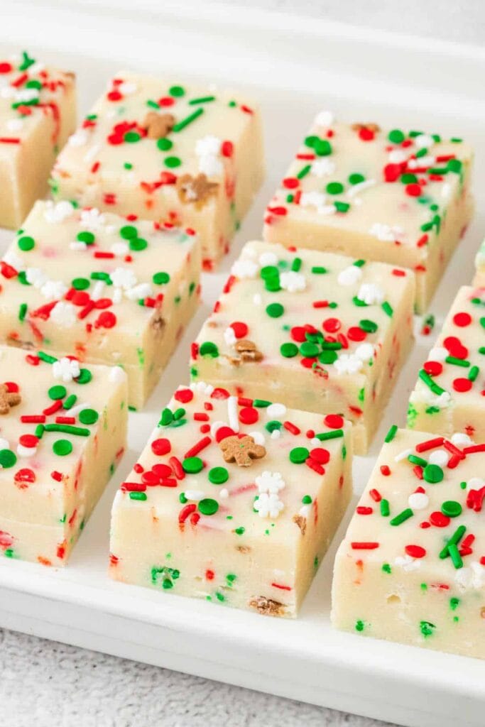 White fudge squares topped with red, green, and holiday-themed sprinkles, neatly arranged on a rectangular tray for a cheerful Christmas treat.