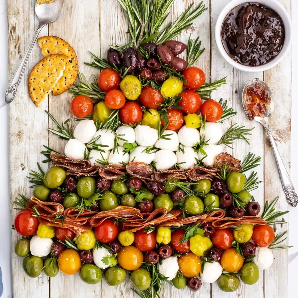 Christmas tree–shaped arrangement of tomatoes, olives, mozzarella, cured meats, and herbs on a wooden surface, with crackers and jam bowl nearby.