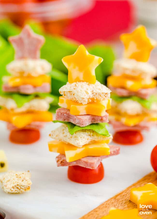 Holiday snack skewers stacked with bread, cheese, meat, and lettuce on cherry tomato bases, topped with star-shaped cheese for a tree-like look.