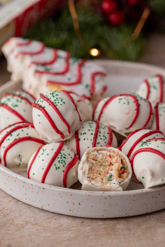 A plate of festive Christmas tree cake truffles coated in white chocolate, decorated with red icing stripes and green sprinkles, with one truffle cut in half to reveal the soft cake filling.