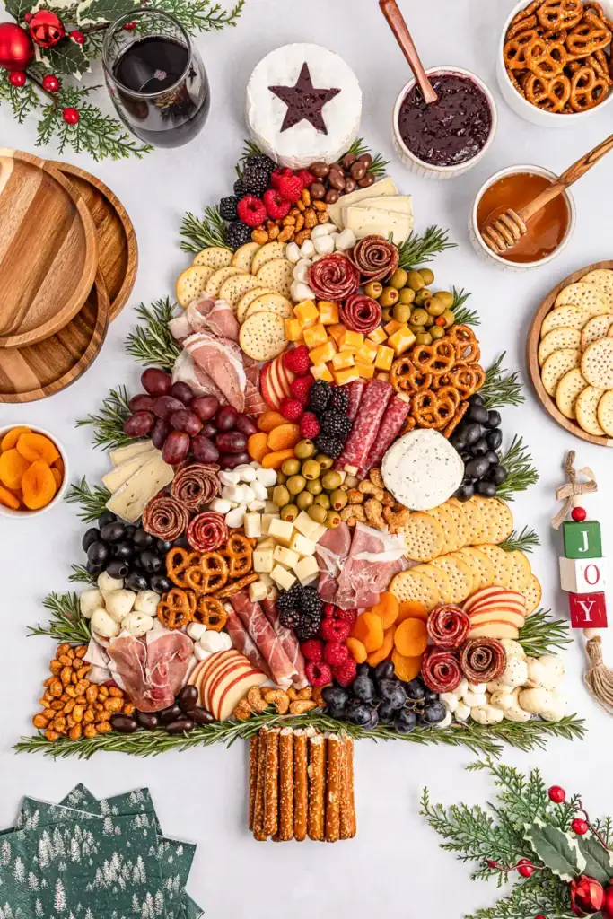 Christmas tree–shaped charcuterie board with layered meats, cheeses, fruits, nuts, crackers, and rosemary sprigs, topped with brie star and pretzel trunk, surrounded by bowls, wine, and holiday decorations.