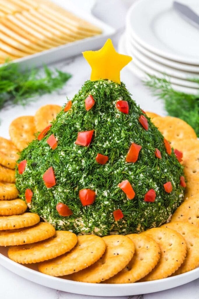 Cone-shaped cheese ball coated in herbs, decorated with red pepper “ornaments” and a yellow star topper, surrounded by crackers on a white plate.
