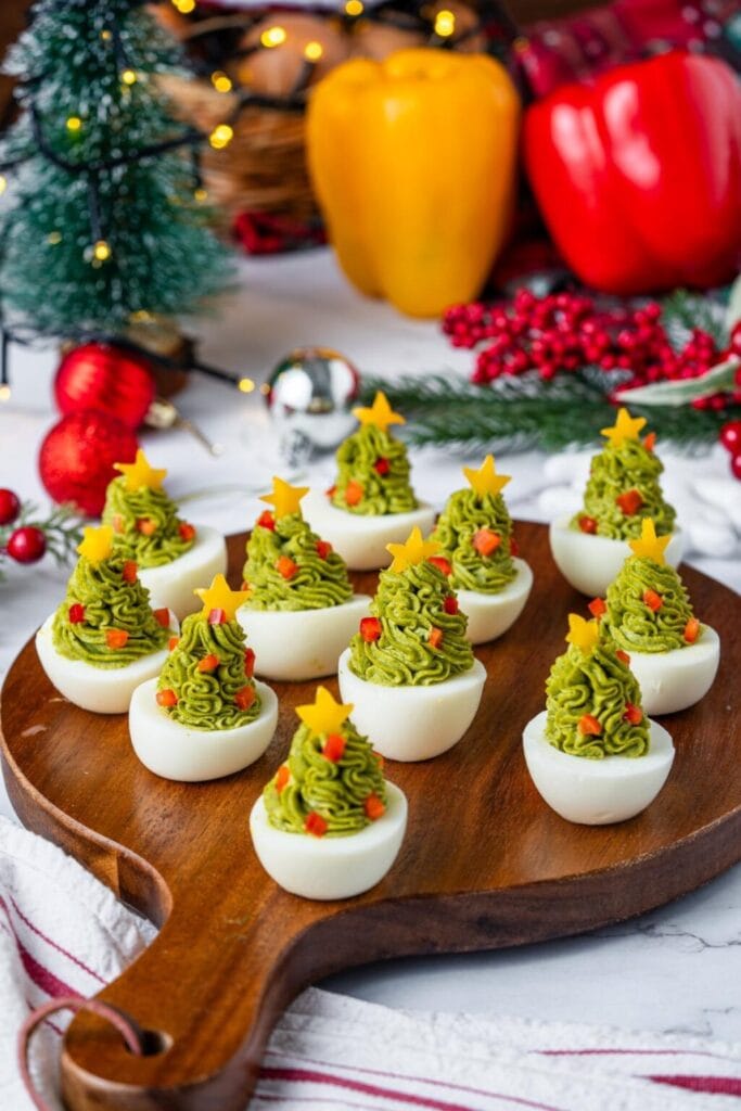 Deviled eggs decorated as mini Christmas trees with green filling, star toppers, and red garnish, arranged on a wooden board with holiday decorations.