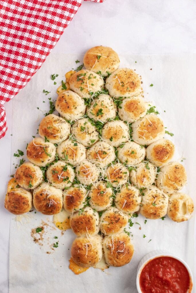 Christmas tree–shaped pull-apart bread rolls sprinkled with parsley and cheese, served on parchment with marinara dipping sauce and checkered cloth nearby.