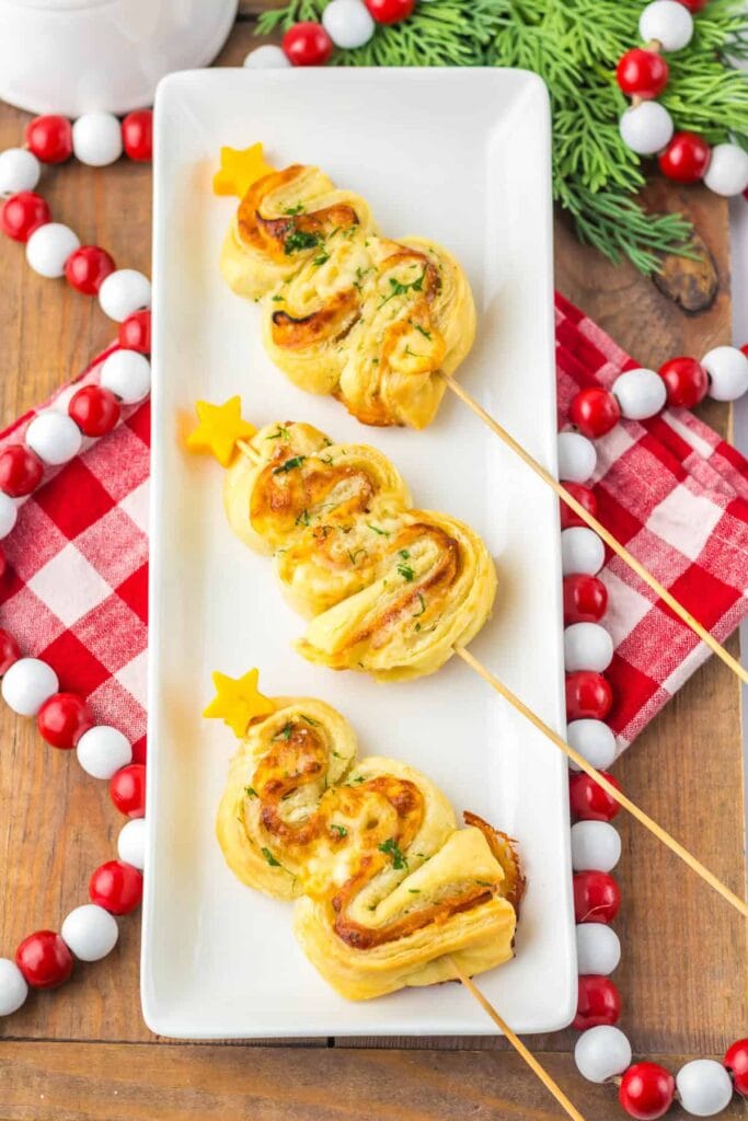 Golden puff pastry Christmas trees on sticks with cheese stars, garnished with herbs, served on a white plate with holiday garland and checkered cloth.