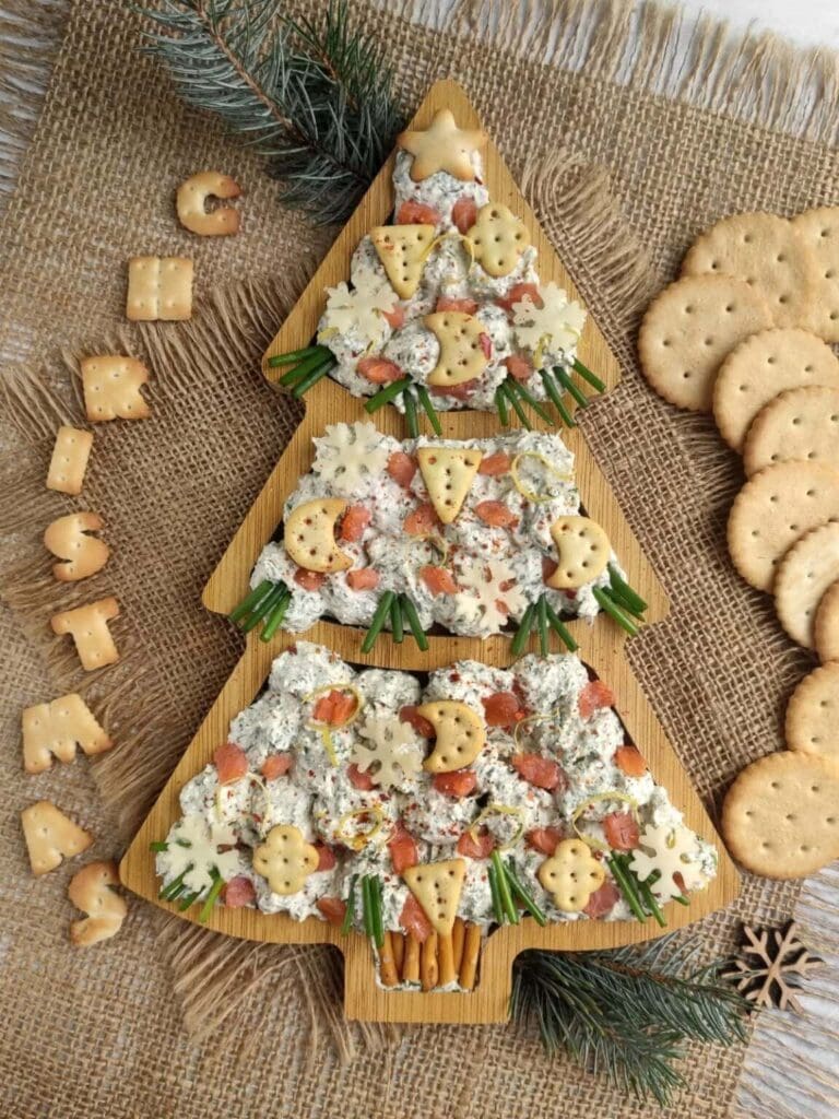 Christmas tree–shaped cheese spread decorated with crackers, pretzels, vegetables, and cookies spelling “CHRISTMAS,” arranged on a wooden board with pine branches.