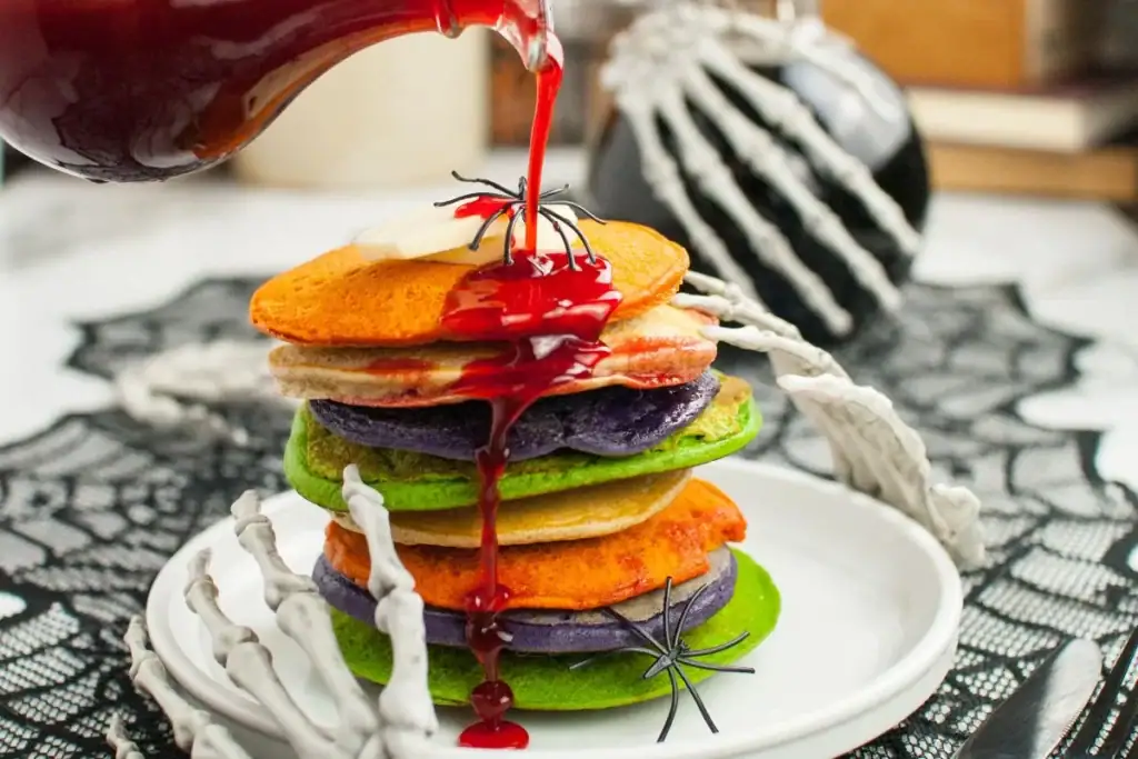 A stack of colorful Halloween pancakes in shades of green, purple, orange, and cream sits on a white plate while bright red syrup is poured over the top. The pancakes are decorated with plastic spiders, and the table is styled with skeleton hand props, black lace, and spooky Halloween decorations in the background.