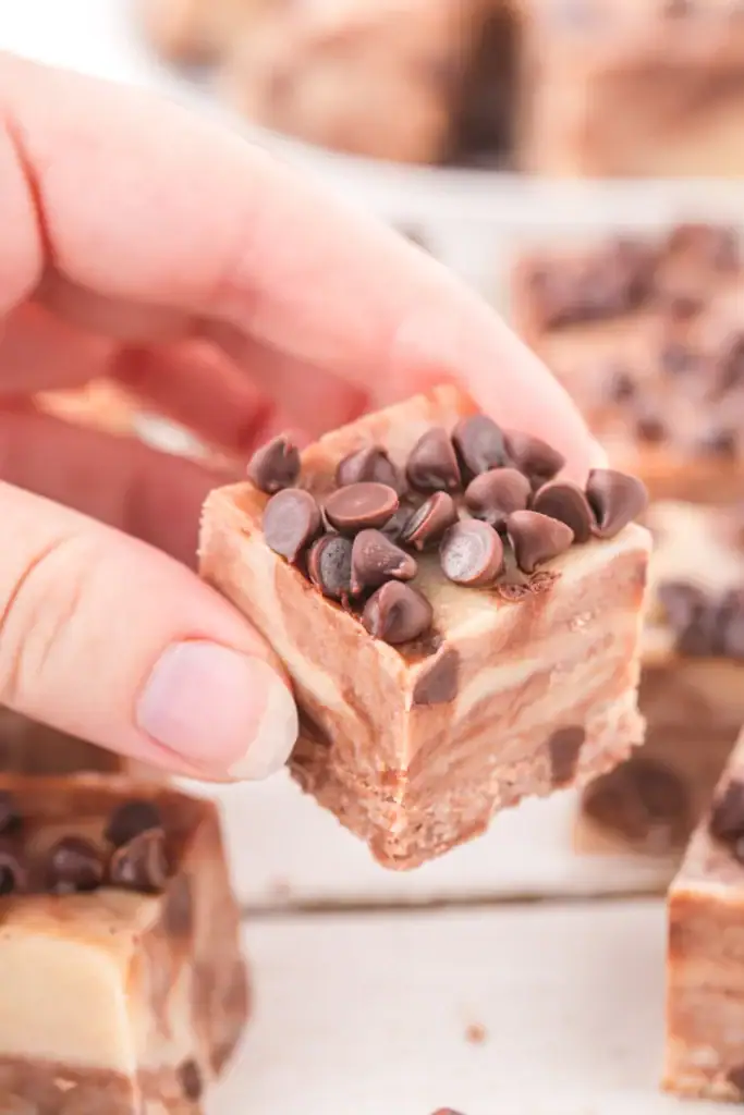 Hand holding layered chocolate fudge square topped with chips, with more stacked pieces in the background.