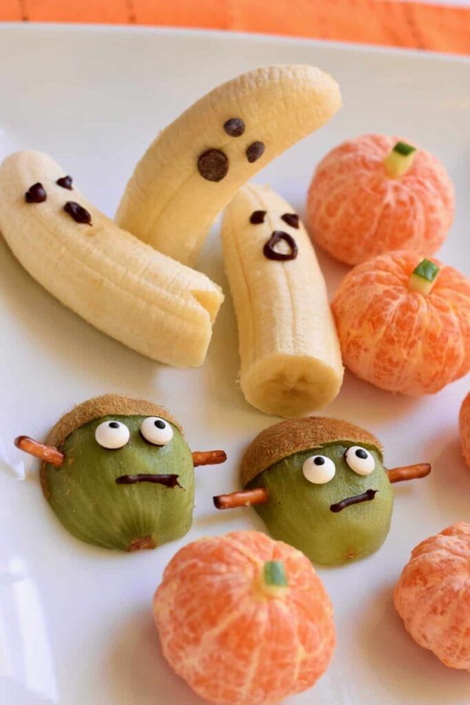 A festive Halloween fruit platter featuring banana ghosts, mandarin orange pumpkins, and kiwi Frankenstein monsters decorated with candy eyes, chocolate, and pretzels for a healthy, spooky snack.