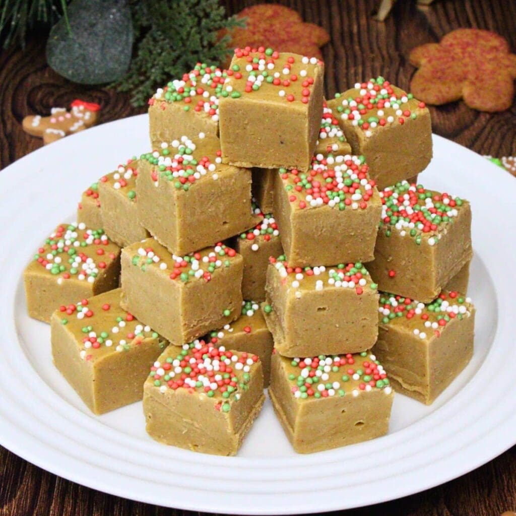 Light brown fudge squares topped with red, white, and green sprinkles, stacked on a white plate with holiday decorations in the background.