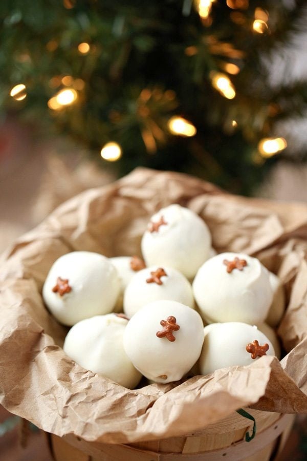 White chocolate-covered Gingerbread OREO truffles topped with tiny gingerbread-shaped sprinkles, arranged in a paper-lined basket with blurred Christmas lights in the background.