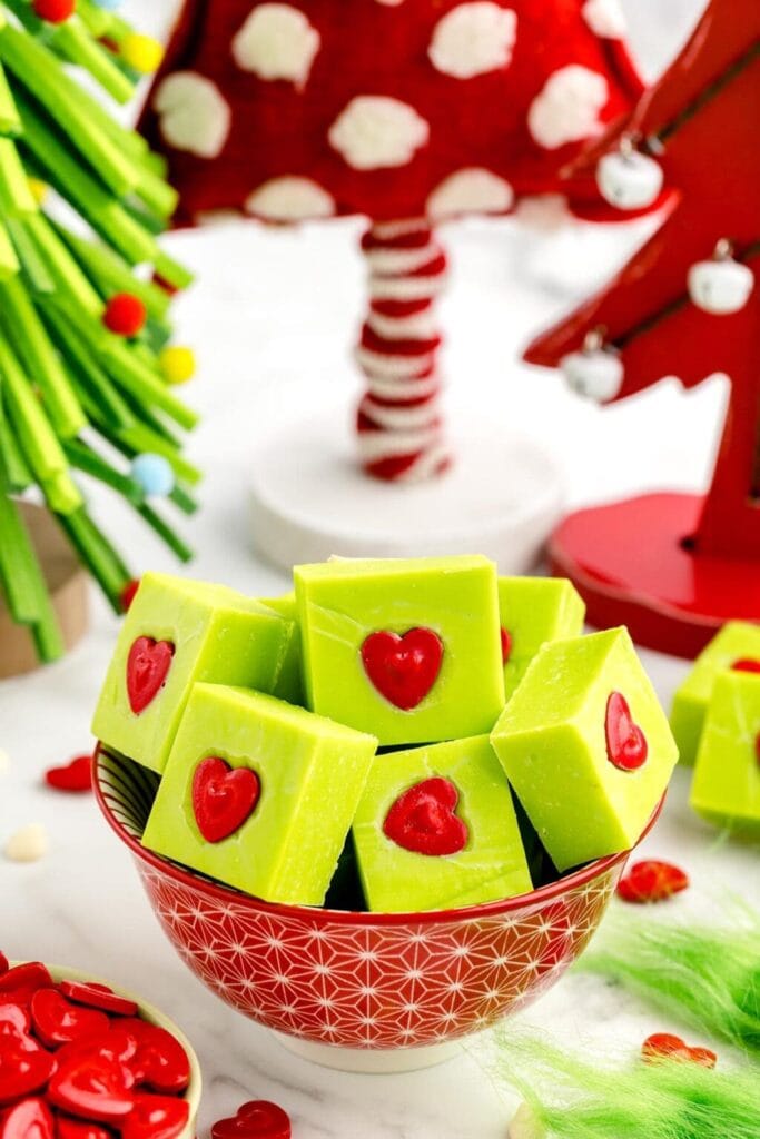 Green square holiday treats with red heart centers in a patterned bowl, surrounded by colorful Christmas decorations.