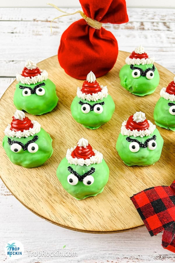 Round wooden board with eight green Grinch-themed Christmas treats decorated with frosting Santa hats, icing eyes, and eyebrows, placed on a white surface with a red bag and plaid fabric nearby.