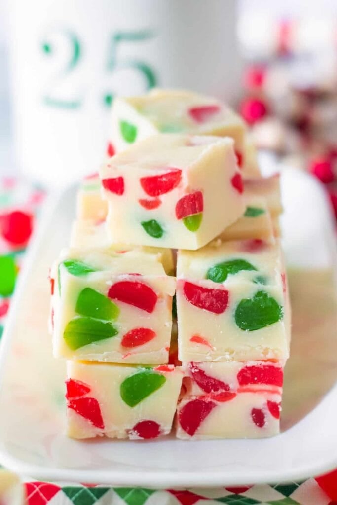 White fudge squares with red and green candied pieces, stacked neatly on a rectangular plate, with blurred holiday décor in background.