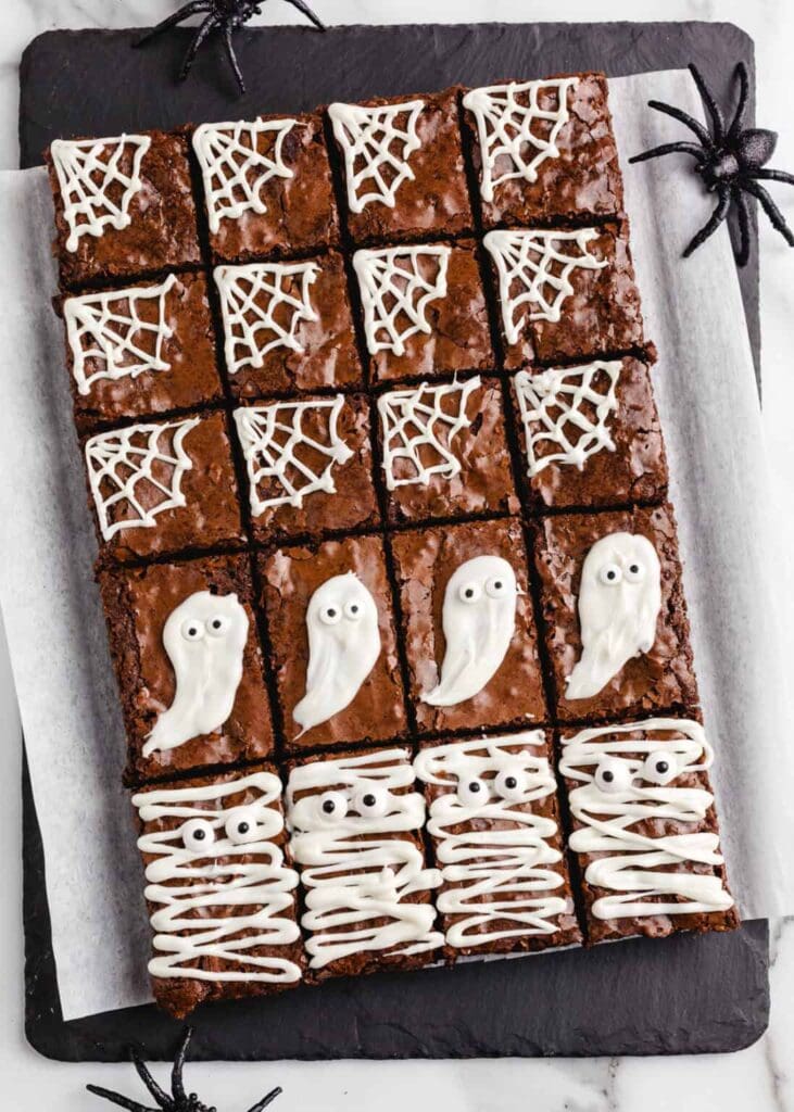 A tray of Halloween brownies is cut into squares and decorated with festive white icing designs. Some brownies feature spiderweb icing, others are topped with ghost-shaped icing decorations, and several are decorated as mummies with white icing bandages and candy eyes. The brownies are displayed on parchment paper over a black serving board, surrounded by plastic spiders for a spooky presentation.