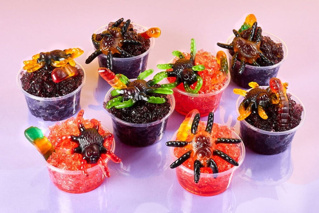 Several small clear cups filled with red and purple Halloween gelatin are topped with gummy spiders, gummy worms, and candy bugs. Some cups are decorated with crushed cookie crumbs to resemble dirt, creating spooky spider nest desserts arranged on a light purple background.