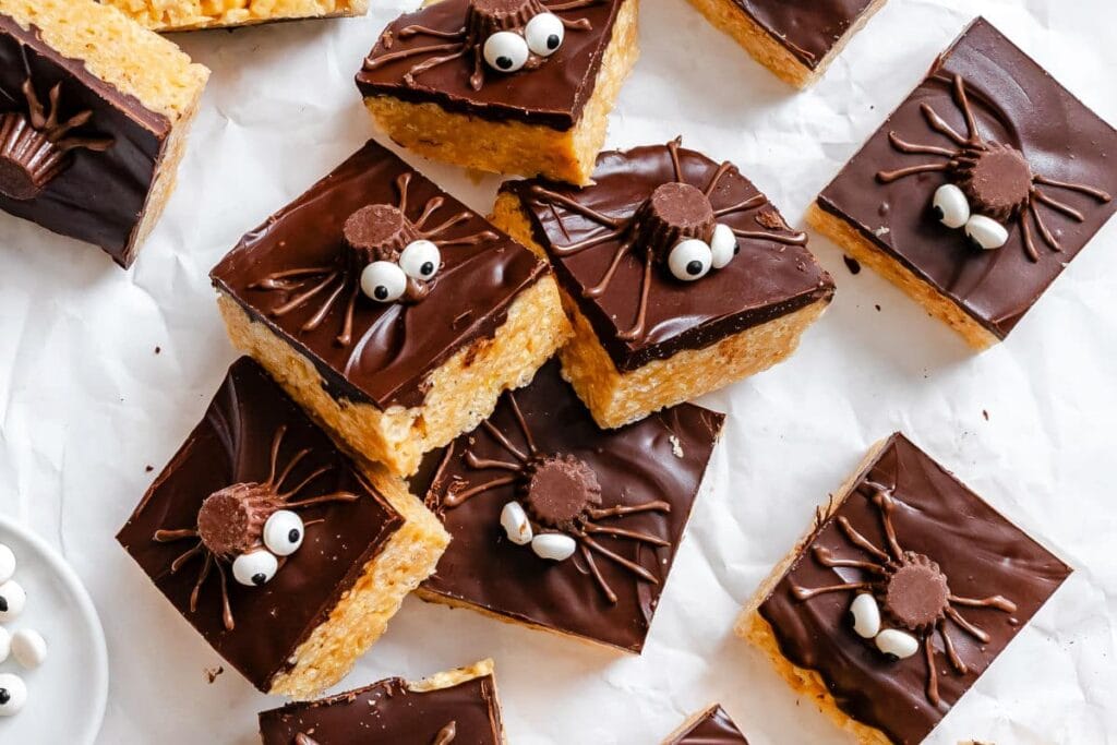 Halloween Rice Krispies treats are cut into squares and topped with a layer of chocolate icing. Each treat is decorated with a candy spider made from a peanut butter cup, candy eyes, and piped chocolate legs. The festive treats are arranged on parchment paper, creating a fun and spooky Halloween dessert display.