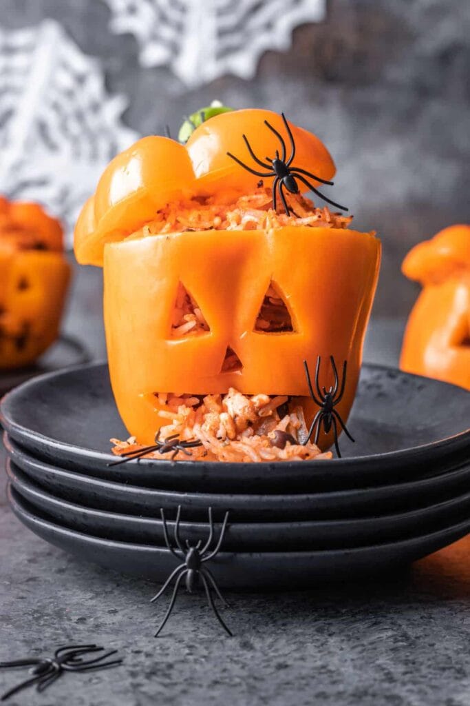 An orange bell pepper carved with a jack-o'-lantern face is stuffed with a rice filling and served on a stack of black plates. The pepper's lid is tilted open, revealing the filling spilling out like a playful Halloween surprise. Plastic spiders decorate the pepper and table, with additional carved peppers and spooky cobweb décor blurred in the background.