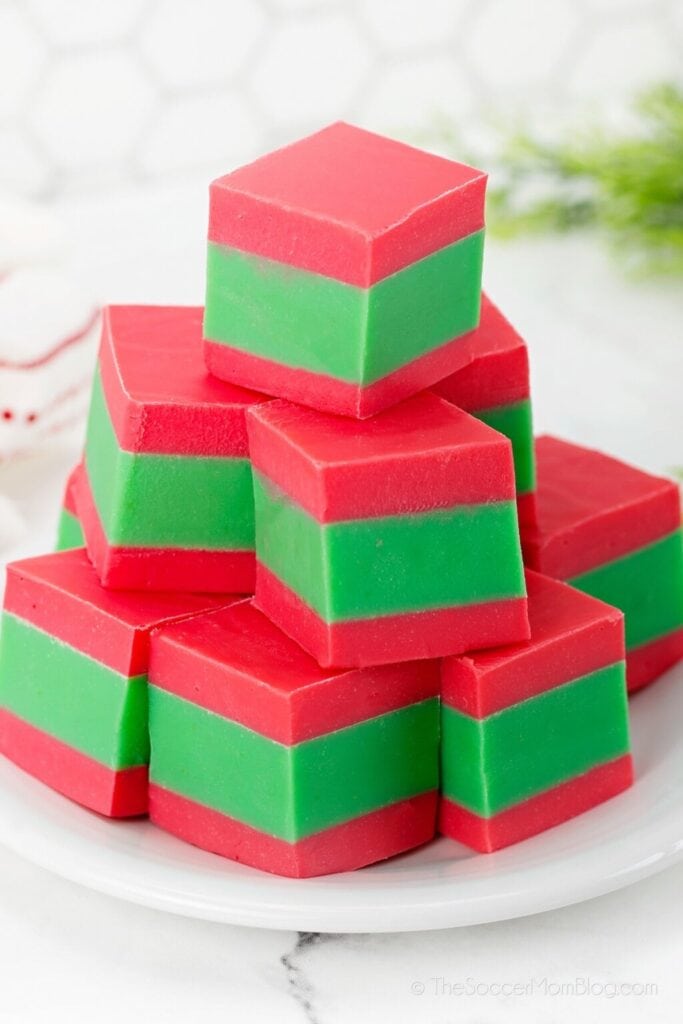 Stacked red and green layered Christmas fudge squares on a white plate, with holiday greenery in the background.