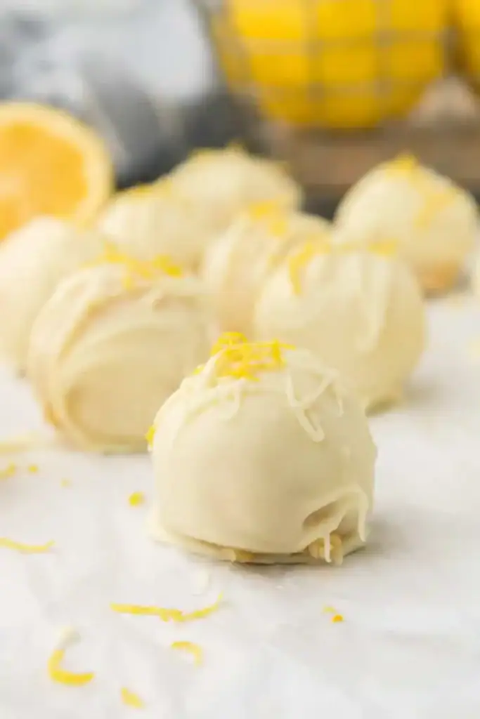 White chocolate-coated lemon truffles topped with fresh lemon zest, arranged on a white surface with lemons blurred in the background.