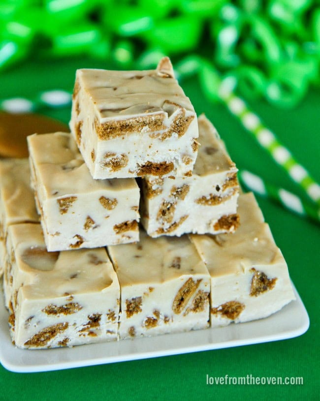 White fudge squares with brown cookie chunks, stacked on a rectangular plate with green festive decorations in the background.