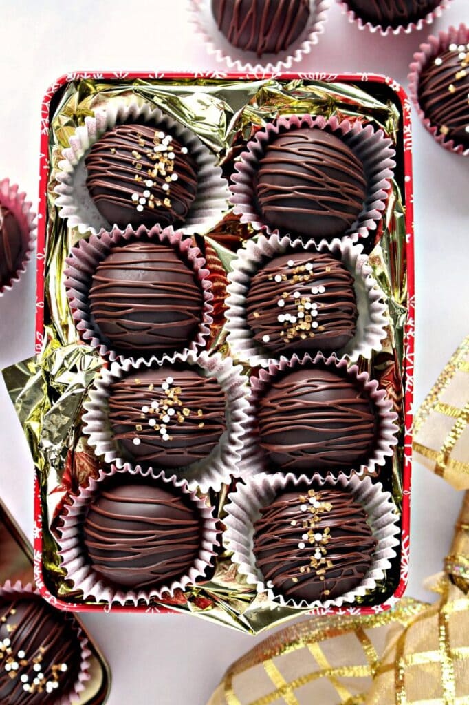 Rectangular tin lined with gold foil, holding nine chocolate truffles in paper cups, decorated with drizzled chocolate and sprinkles, with extra truffles and a gold ribbon nearby.