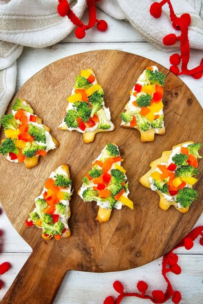 Christmas tree–shaped pastry snacks topped with cream cheese, broccoli, and colorful bell pepper pieces, arranged on a wooden board with holiday garlands.