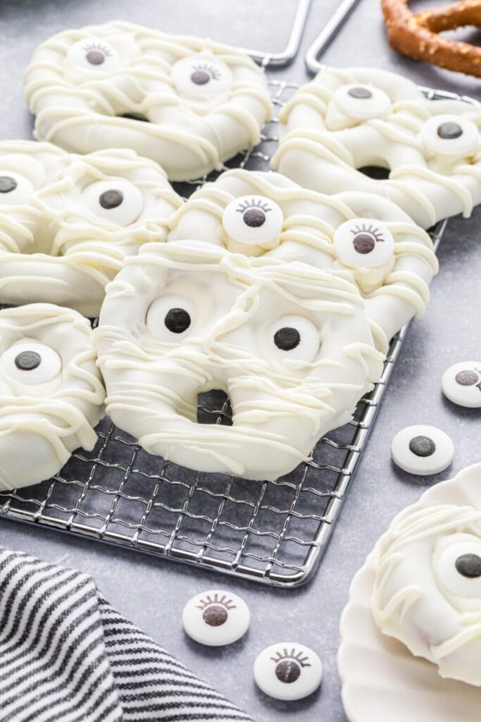 Several Halloween mummy pretzels are coated in white chocolate and decorated with candy eyes and white chocolate drizzle to resemble wrapped bandages. The spooky treats are arranged on a cooling rack, with extra candy eyes scattered around and a pretzel visible in the background.