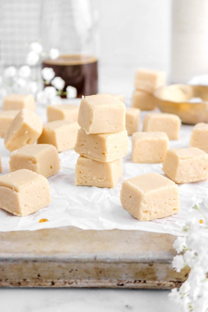 Light beige fudge squares with smooth texture, stacked on parchment-lined tray beside blurred coffee glass and white flowers.