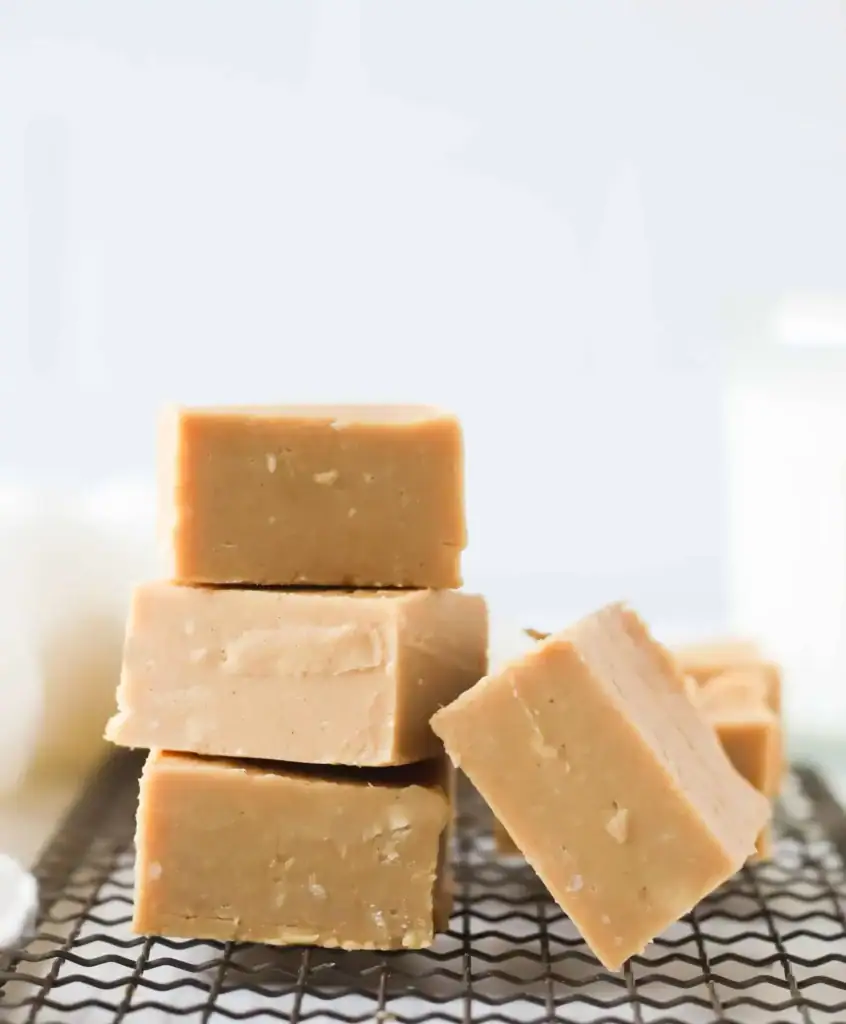 Light brown fudge cubes stacked on a cooling rack, with smooth surfaces and crumbly edges highlighted against a soft background.