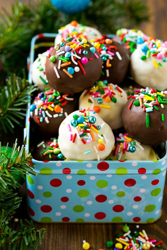 Chocolate and white chocolate-coated Oreo balls topped with colorful sprinkles, arranged in a festive polka-dot tin surrounded by holiday greenery.