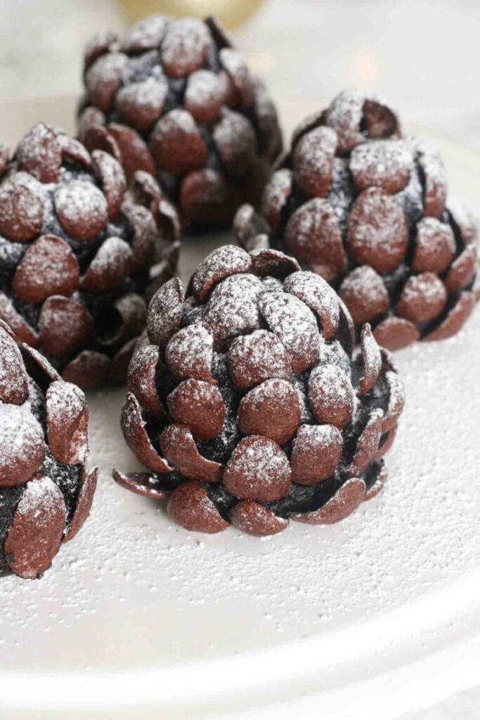 Chocolate Oreo pinecone truffles coated with chocolate cereal pieces and dusted with powdered sugar, arranged on a white serving plate.