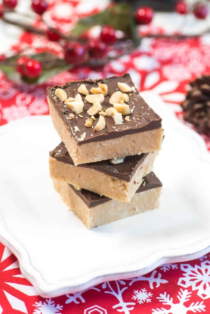 Stacked chocolate-topped peanut butter bars with chopped nuts on a white plate, set against a red snowflake tablecloth with pinecones and berries.
