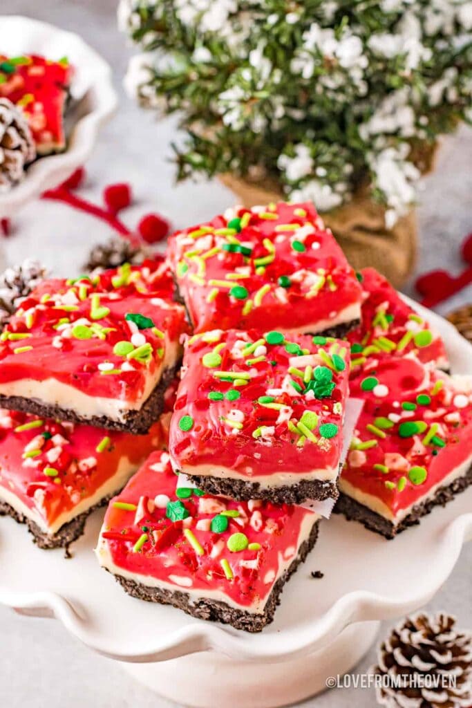 Layered holiday bars with chocolate crust, creamy middle, and red sprinkle-topped layer, stacked on a white cake stand with pinecones and Christmas accents.