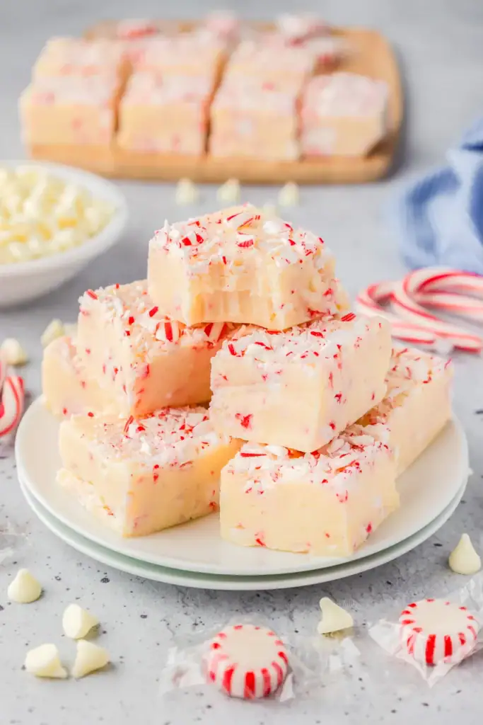 White peppermint fudge squares with crushed candy topping, stacked on a plate with candy canes, chocolate chips, and festive props nearby.