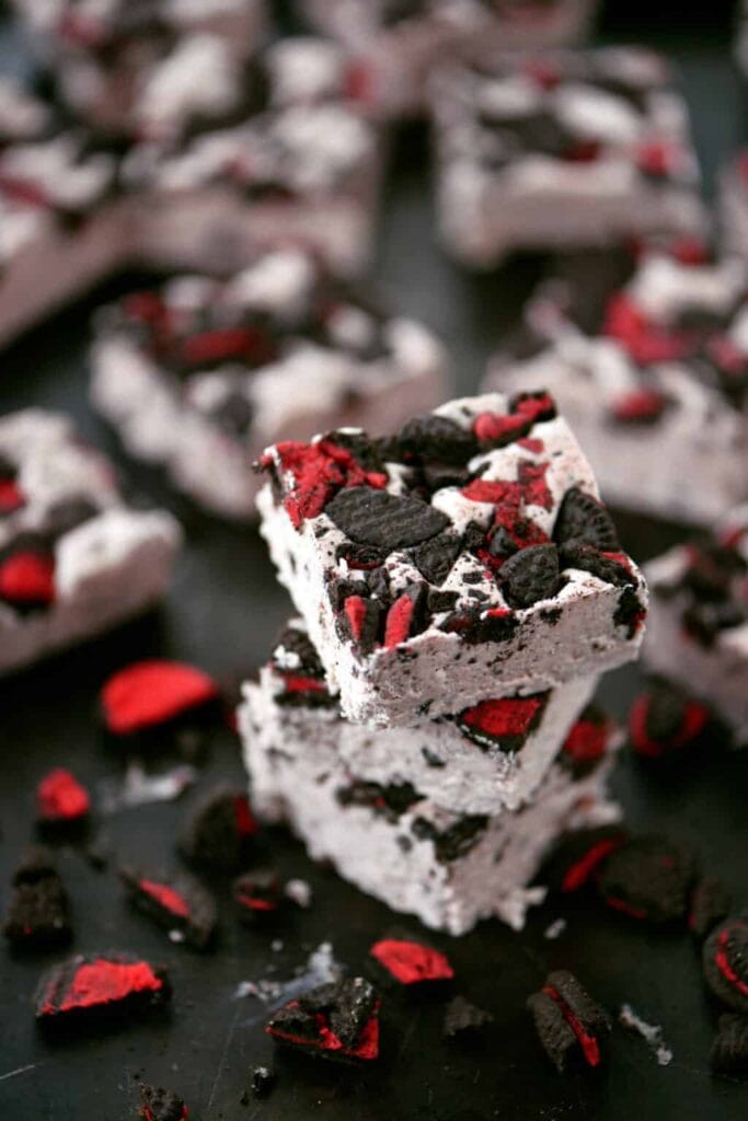 White fudge squares with chunks of black and red cookies, stacked on a dark surface for a bold holiday look.