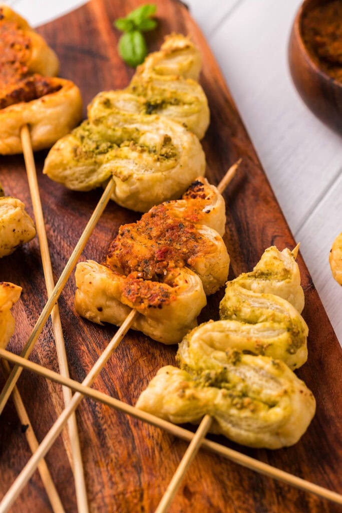 Golden puff pastry twists on skewers with pesto and tomato fillings, arranged on a wooden board beside basil and dipping sauce.