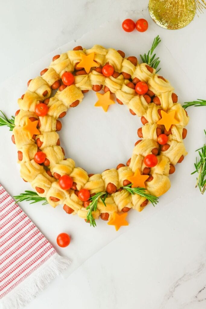 Wreath-shaped arrangement of crescent-wrapped sausages decorated with rosemary, cherry tomatoes, and cheddar stars on a white surface with festive cloth nearby.