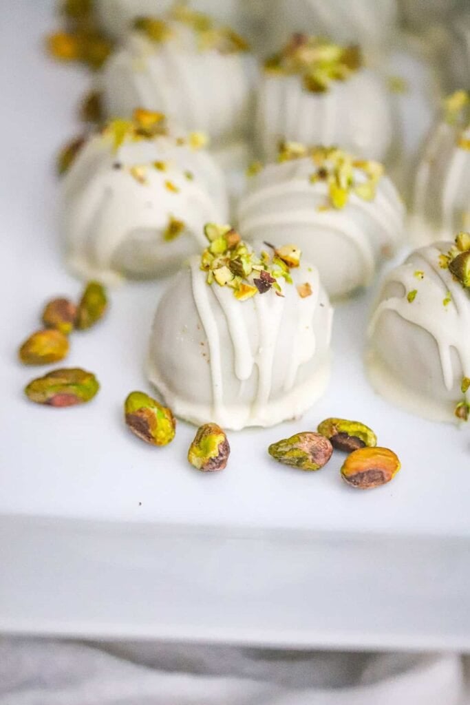 White chocolate-coated pistachio cookie truffles drizzled with white chocolate and topped with chopped pistachios, arranged on a white serving platter.