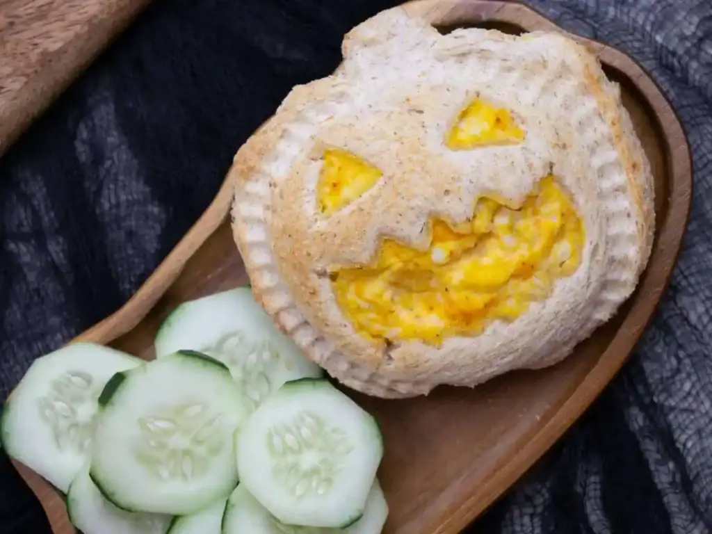 A Halloween-themed egg sandwich is served on a wooden tray, with toasted bread cut into a smiling jack-o'-lantern face that reveals fluffy scrambled eggs through the carved eyes and mouth. Fresh cucumber slices are arranged beside the sandwich, with a dark cloth backdrop adding to the festive Halloween presentation.