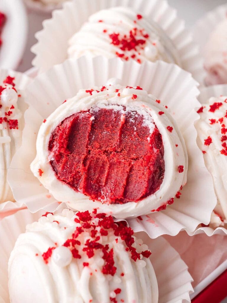 White chocolate-coated red velvet truffles decorated with red cake crumbs and white sprinkles, with one truffle cut open to reveal the rich red velvet cake center.