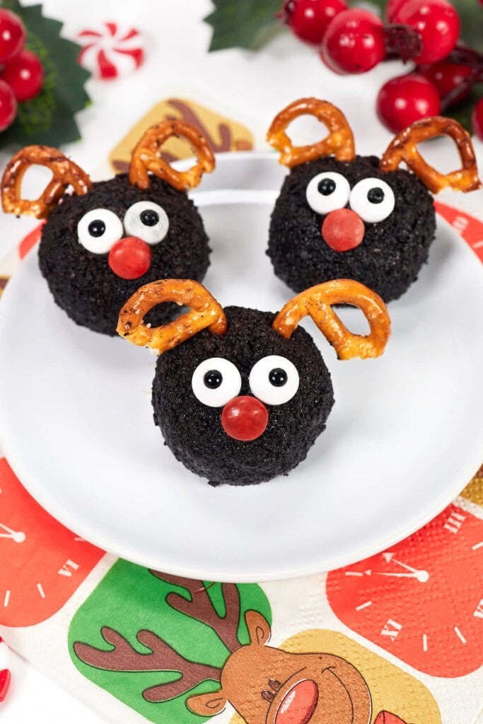 Three chocolate reindeer treats with candy eyes, red noses, and pretzel antlers on a white plate over a festive napkin, with berries and peppermint candy in the background.