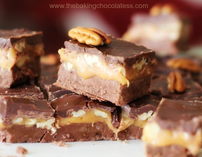 Square chocolate caramel fudge bars with pecan topping, showing rich chocolate and gooey caramel layers stacked on a white surface.