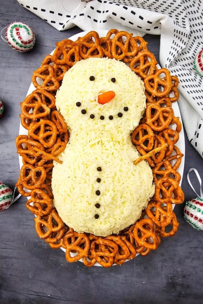 Snowman-shaped cheese ball with peppercorn eyes, carrot nose, and pretzel buttons, surrounded by pretzels on a white platter with festive decorations.