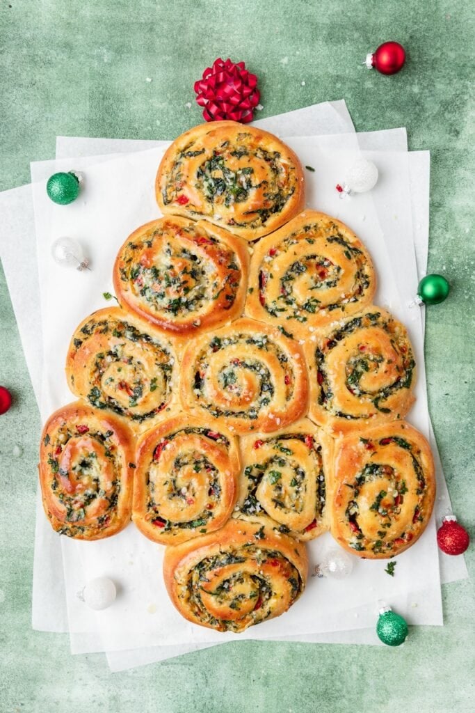 Christmas tree–shaped spiral bread rolls filled with herbs, cheese, and peppers, arranged on parchment with holiday ornaments and red bow topper.