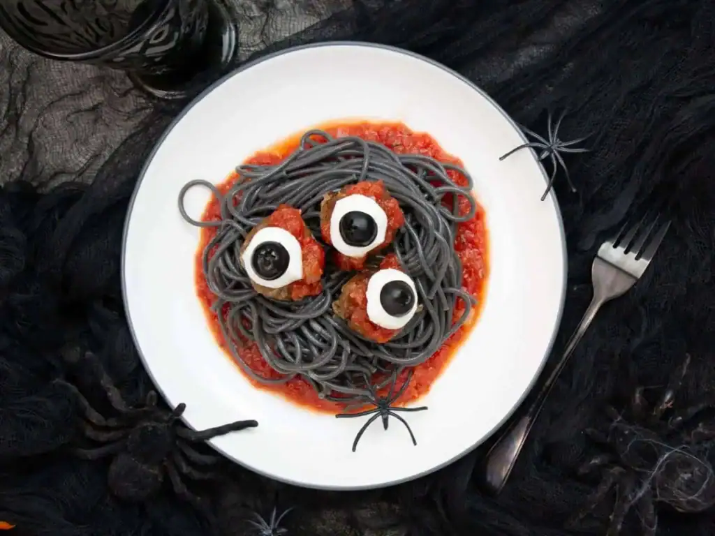 A Halloween-themed plate of black spaghetti is served over red tomato sauce and topped with three meatballs decorated as eyeballs using mozzarella slices and black olive centers. The dish is styled with black fabric, plastic spiders, and spooky table décor, creating a creepy yet festive Halloween meal.
