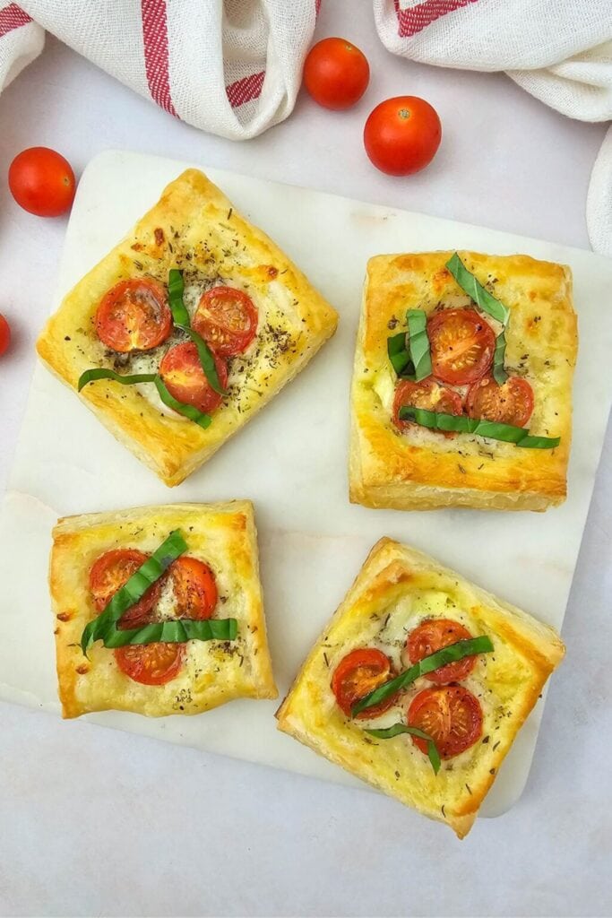 Square puff pastry tarts topped with cherry tomatoes, basil, melted cheese, and herbs, arranged on a white surface with scattered tomatoes and striped cloth.
